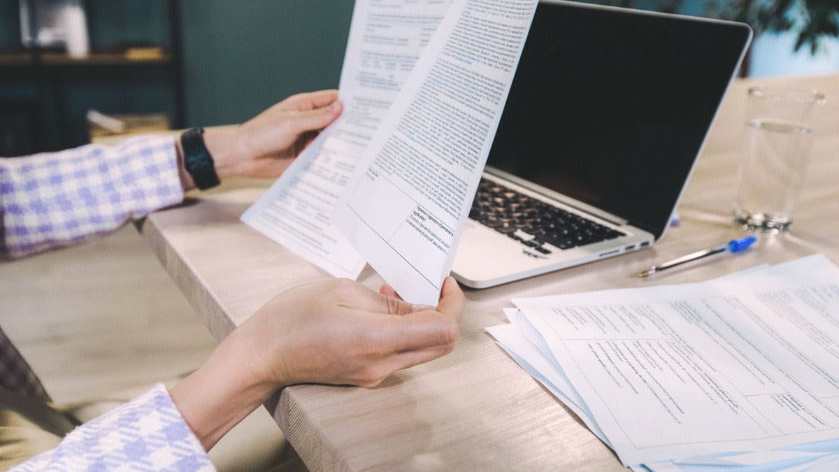 Hands hold a printed multi-page document above a wooden desk next to an open laptop.