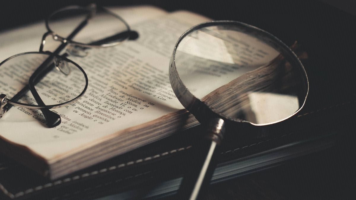 A magnifying glass rests over an open book's dense text beside a pair of reading glasses.