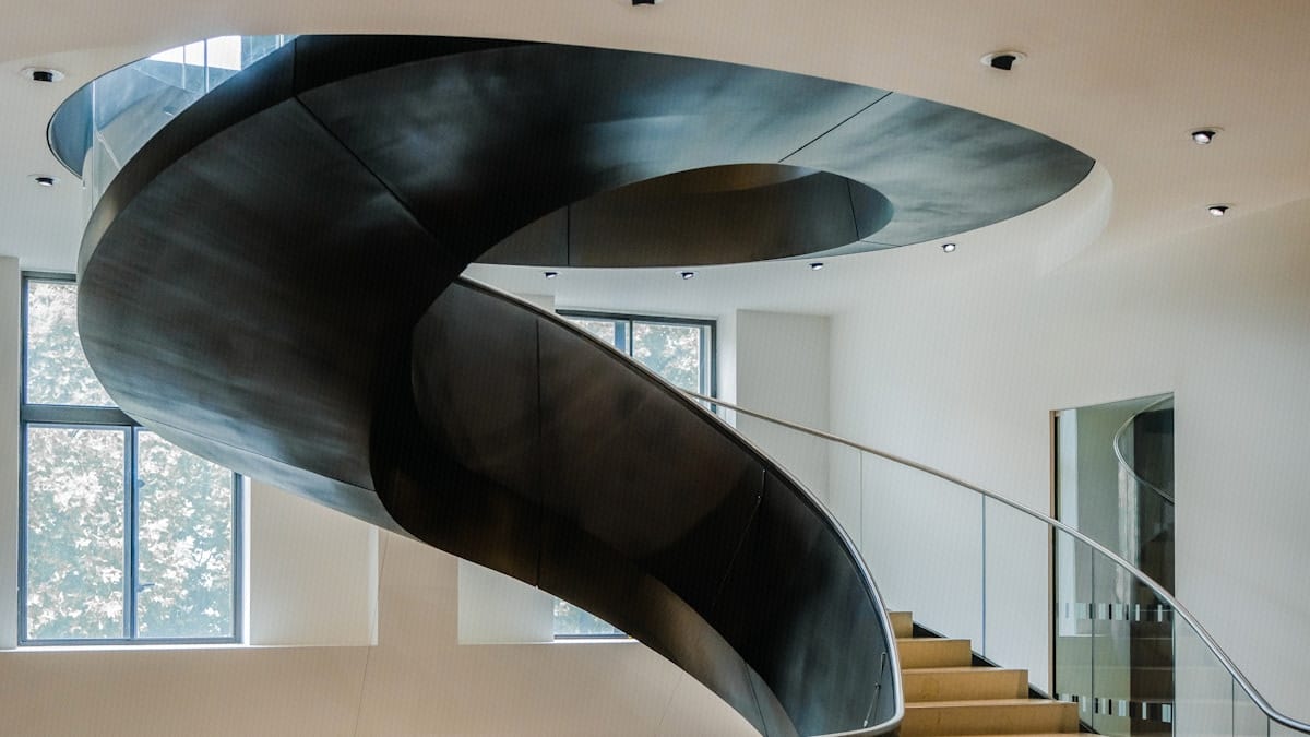 A sweeping modern spiral staircase curves upward through a light-filled interior, viewed from below.