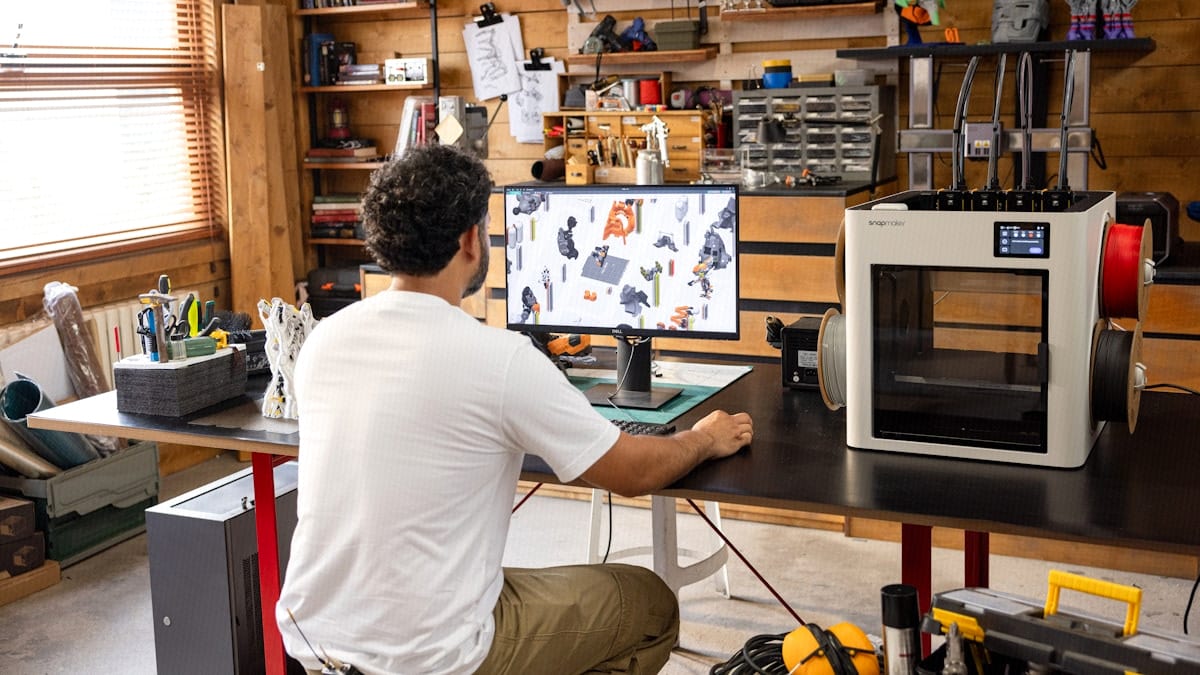 A maker works from behind at a monitor showing a CAD design, next to a 3D printer loaded with filament in a home workshop.