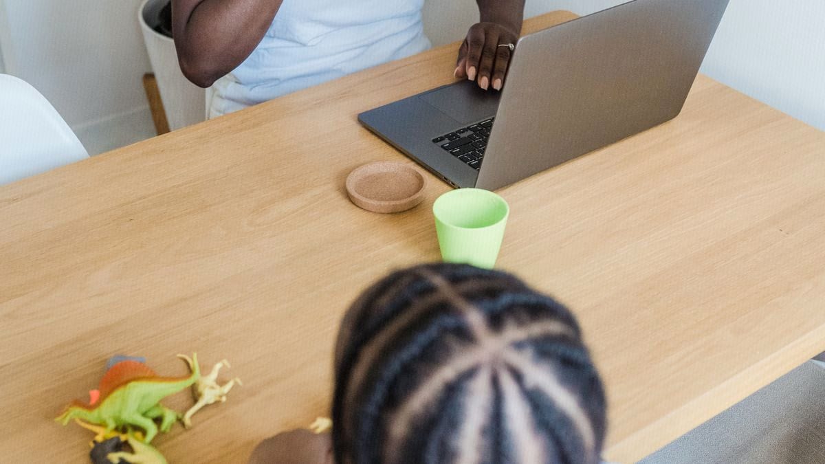 A parent works on a laptop at a wooden table while a child plays quietly nearby with toy dinosaurs.