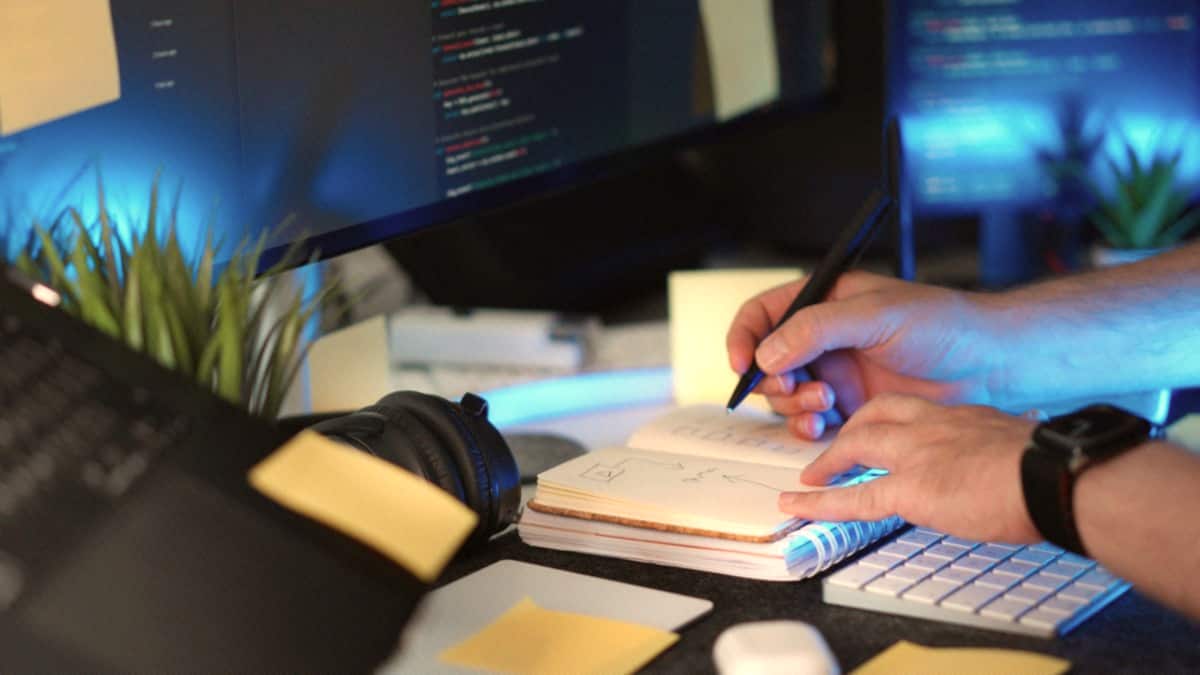Hands write notes in a notebook beside a keyboard and glowing monitors on a warm desk.