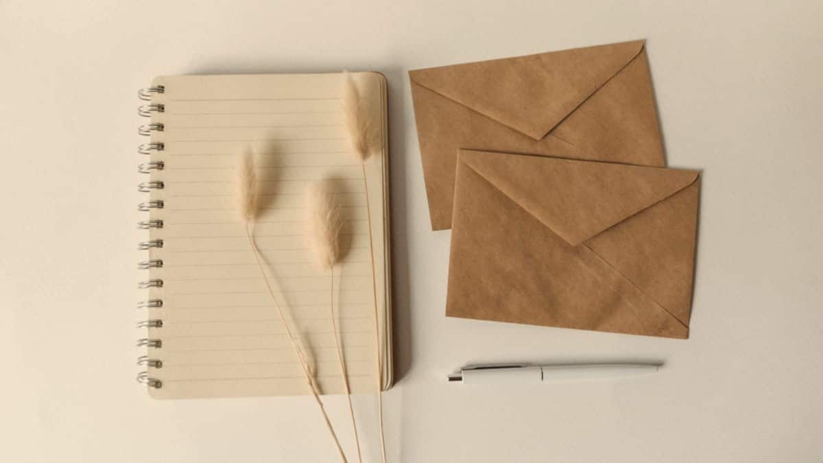 A spiral notebook, dried grass stems, kraft envelopes, and a pen are arranged on a warm off-white desk.