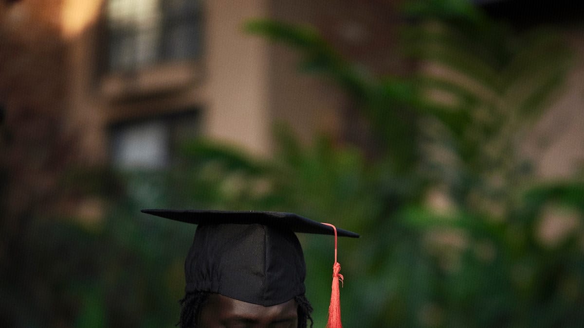 A graduate wears a mortarboard cap with a red tassel, softly lit against a green campus backdrop.