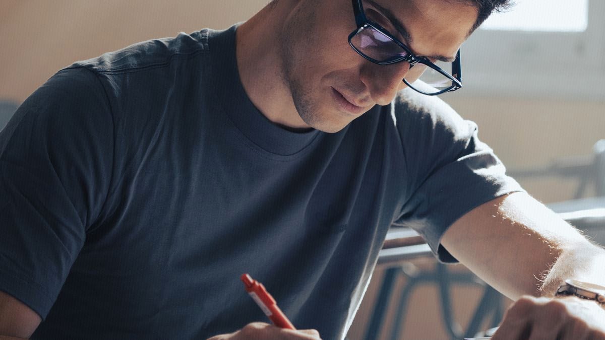 A young man in glasses concentrates while writing during a proctored exam, sunlight from a window behind him.